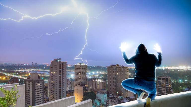 The silhouette of a shrouded figure perches on a skyscrapers ledge. His hands are filled with light and held above his head, as off in the distance two bolts of lightning form, mirroring the position he has made.