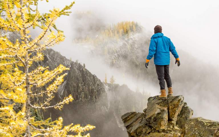 Travel Photos. Featured LIFELIGHTLENS image of a man standing on a cliff looking out on larches in the fog.